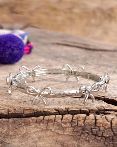 The Bow Bangle Bracelet - Silver Tone features a hammered texture and small, evenly spaced bow-shaped charms. It rests on a rustic wooden surface with a blurred purple bead accessory in the background.