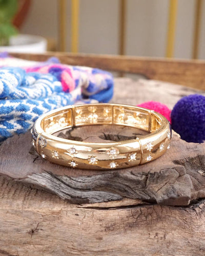 The Wide Stretch Bangle with Clear Crystals – Gold-Tone rests on wood, surrounded by colorful fabric and round, textured accents in the background.