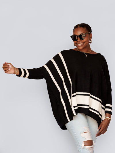 A person smiles, wearing the Lydia Striped Batwing Sweater in black with a relaxed silhouette, poses with one arm extended. They have braided hair, hoop earrings, and are dressed in ripped light blue jeans against a plain background.