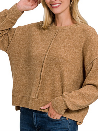 A person wearing the Cassidy Stitch Detail Top in Deep Camel paired with blue jeans is smiling, gently touching their hair. The loose-fitting sweatshirt features a central seam design and a high-low hem.