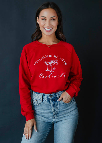 A woman smiles brightly in a Beginning To Look A Lot Like Cocktails Sweatshirt - Red, featuring playful text and three cocktail glass images. Her long dark hair complements her blue jeans against a dusky background, dressed in comfort with polyester cotton spandex.