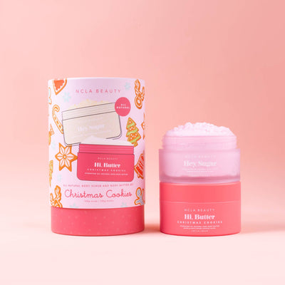 A pink NCLA Beauty Christmas Cookies Body Scrub + Body Butter Holiday Gift Set sits against a soft pink background. The collection showcases a "Hi, Butter" body butter jar alongside a matching cylindrical container decorated with illustrations of holiday cookies, evoking a festive and cozy atmosphere.