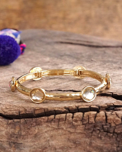 The Faceted Glass Golden Bangle - Yellow features a hammered gold-tone band with five round, clear gemstones and is shown on a rustic wood surface with a blurred purple and blue accent in the background.