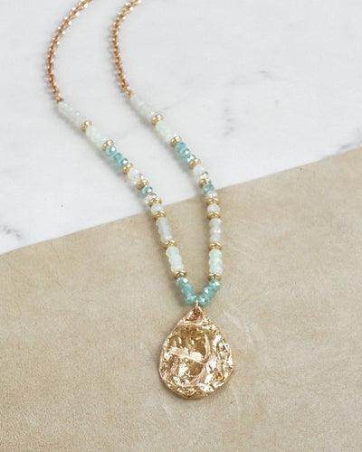 The Long Beaded Necklace with Golden Teardrop Pendant - Green features light blue, mint aqua, and white beads with gold accents and a textured gold teardrop pendant, shown on a beige and marble backdrop.