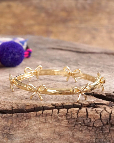 The Bow Bangle Bracelet - Gold features multiple small bow-shaped designs with a stretchy fit, shown on a rustic wooden surface alongside a purple decorative item in the background.