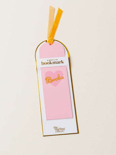 A pink acrylic bookmark with a heart and the word 'Books' in gold, packaged with a yellow ribbon at the top. The bookmark is labeled 'acrylic bookmark' and branded by 'the darling effect.