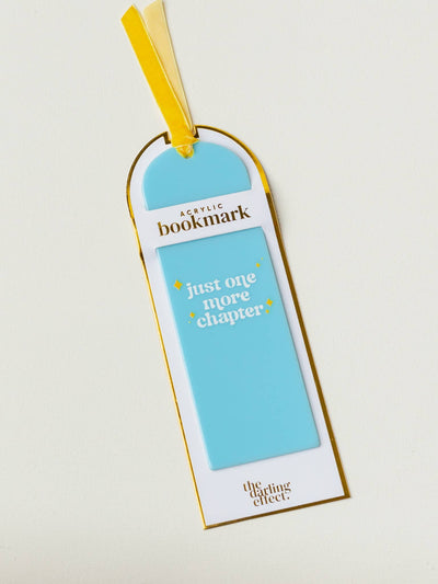 A blue acrylic bookmark with the phrase "just one more chapter" in white lettering, accented with small yellow diamond shapes. The bookmark is displayed in its packaging with a yellow ribbon attached.