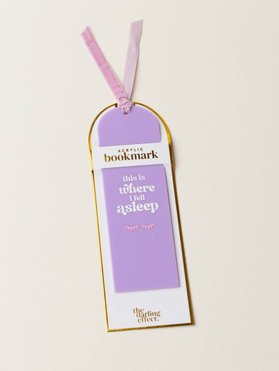 A lavender acrylic bookmark in its packaging, featuring the phrase "this is where I fell asleep" in white text with a closed-eye illustration. The packaging includes a gold trim and label reading "Acrylic Bookmark," with a soft purple ribbon tied at the top.