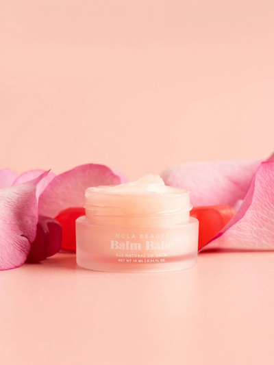 An open small jar of Balm Babe Candy Roses Lip Balm reveals its natural contents, surrounded by soft pink and red rose petals on a pink background.