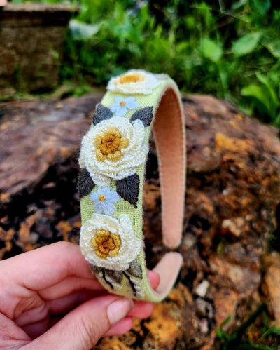 A hand holds the Floral Embroidered Green Headband - Cream Yellow, adorned with hand-stitched white and yellow flowers and green leaves, set against a blurred outdoor background with grass and a tree stump.