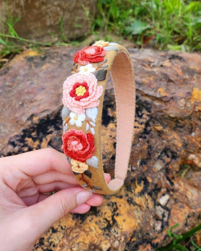 A hand holds the Floral Embroidered Mustard Headband - Peach, featuring colorful hand-stitched flowers in red, pink, yellow, and white. Shown outdoors on a rock with grass—this boho accessory adds charm to any outfit.
