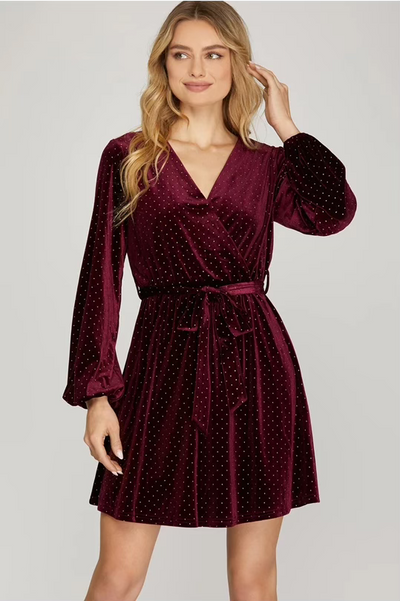 A woman is wearing the Harper Surplice Dress in wine red, which has long sleeves and exquisite gold dot detailing. This velvet wrap dress features a tied waist. She stands against a plain background, smiling and touching her hair.