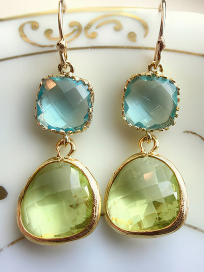 A pair of elegant earrings featuring 13mm faceted peridot teardrop gems paired with 9mm aquamarine square blocks, both set in gold-plated bezels with 14k gold-filled earwires. Displayed on a white ceramic dish with gold detailing, these earrings are dainty and versatile.