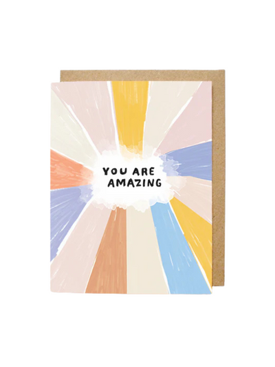 A greeting card with a colorful abstract design featuring pastel rays in shades of orange, yellow, pink, blue, and cream radiating outward. In the center, a white cloud shape contains the bold, black text "YOU ARE AMAZING." The card is paired with a brown kraft envelope in the background.