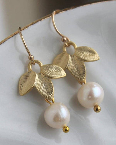The Freshwater Pearl Leaf Earrings - Gold feature textured gold leaf designs with dangling round freshwater pearls, displayed on a white ceramic surface for an elegant look suitable for any occasion.