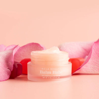 A small, round jar of Balm Babe Candy Roses Lip Balm from NCLA Beauty is set against a soft pink background, surrounded by pink rose petals. The vegan lip balm boasts a creamy texture, with the label on the jar clearly visible.