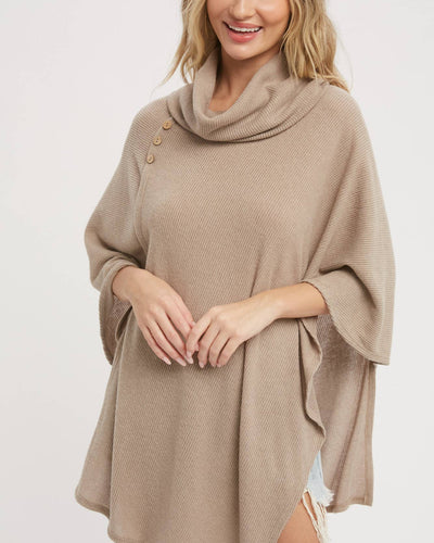 A woman with wavy blonde hair smiles with hands clasped, wearing the Asymmetrical Cowl Neck Poncho in Latte—a lightweight, beige ribbed knit featuring a cowl neck and three shoulder buttons that drapes loosely for effortless style.